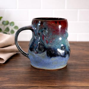 Op de afbeelding: Een handgemaakte keramische mok met een unieke, organische vorm. De mok heeft een mix van kleuren, waaronder diep rood, blauwgroen en blauw, met een donker handvat. De mok is ongeveer 10 cm hoog en is geschikt voor warme of koude dranken.