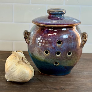 Op de afbeelding: Een keramische knoflookhouder met deksel, in blauw, paars en bruin. De houder heeft ventilatiegaten en handvatten. Er ligt een hele knoflookbol naast. De houder staat op een houten oppervlak.