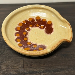 Op de afbeelding: Een keramische lepelhouder met een lichtgele basis en een bruin en oranje abstract ontwerp. De lepelhouder heeft een gebogen vorm met een kleine lip aan één kant.