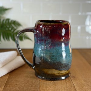 May include: A handmade ceramic mug with a large handle. The mug features a gradient of colors, including deep red at the top, transitioning to teal and brown at the base. The mug is on a wooden surface.