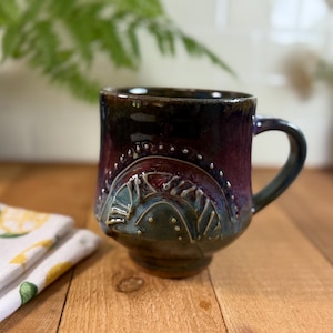 Puede incluir: Una taza de cerámica con un esmalte oscuro y brillante que transiciona de un violeta profundo en el borde a un tono azul verdoso y marrón en la base. El exterior presenta un patrón de arco en relieve decorado con pequeños puntos espaciados.