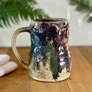 May include: A handmade ceramic mug with a unique textured surface. The mug features a handle and is adorned with a blend of colors, including blue, green, and burgundy. The mug is approximately 5 inches tall.
