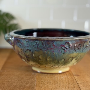 Op de afbeelding: Een handgemaakte keramische kom met een unieke glazuur. De kom heeft een teal, paars en geel kleurenschema met hartvormige ontwerpen rond de rand. Het heeft twee kleine handvatten en een stevige basis, ideaal om te serveren of te presenteren.