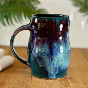 May include: A handmade ceramic mug with a unique glaze. The mug features a dark teal base that transitions to blue and burgundy hues. The mug has a large handle and is approximately 5 inches tall.