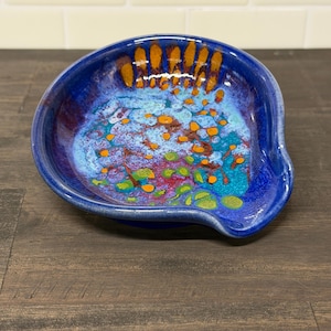 May include: A handmade ceramic spoon rest with a vibrant, abstract design. The spoon rest features a deep blue exterior with a colorful interior of turquoise, orange, and green accents. The unique shape and glaze give it a handcrafted feel.