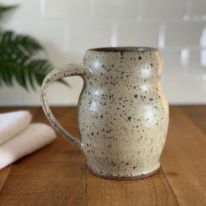 Op de afbeelding: Een bespikkelde keramische mok met een afgeronde body en een grote handgreep. De mok is beige met donkerbruine spikkels en een bruine rand. De mok staat op een houten oppervlak.