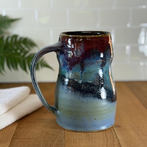 May include: A handmade ceramic mug with a large handle. The mug features a blend of colors, including blue, green, and brown, with a glossy finish. The mug is approximately 5 inches tall and is suitable for hot or cold beverages.