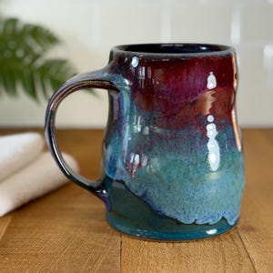 May include: A handmade ceramic mug with a glossy finish. The mug features a gradient of colors, transitioning from deep red at the top to teal and blue at the bottom. It has a large handle and a wide opening, perfect for coffee or tea.