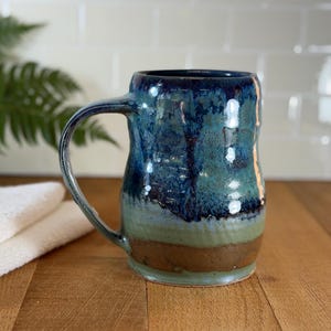 May include: A handmade ceramic mug with a large handle. The mug features a blue and green glaze over a brown base. The mug is approximately 5 inches tall and is sitting on a wooden surface.