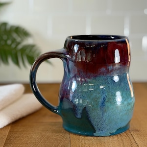 May include: A handmade ceramic mug with a unique glaze. The mug features a gradient of colors, including deep red, purple, and teal. The handle is a dark blue. This stoneware mug is perfect for coffee or tea.