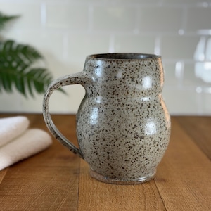 Op de afbeelding: Een set van drie keramische mokken met een gespikkelde, matte afwerking. De mokken zijn in crème, lichtgrijs en donkergrijs. Elke mok heeft een eenvoudig, minimalistisch ontwerp en een afgeronde handgreep.