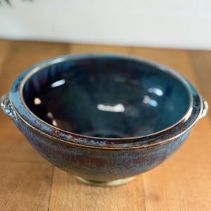Op de afbeelding: Een handgemaakte keramische kom met een glanzend, donkerblauw glazuur. De kom heeft een brede rand met een lichtere blauwe rand en kleine handvatten. De buitenkant heeft een gevlekte, aardse bruin-blauwe kleur.