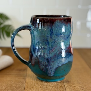 May include: A handmade ceramic mug with a unique, organic shape. The mug features a blend of teal, blue, and maroon glazes, creating a marbled effect. The mug has a large handle and is suitable for hot or cold beverages.