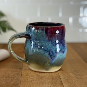 May include: A handmade ceramic mug with a unique glaze. The mug features a blend of colors, including teal, blue, and burgundy, with a glossy finish. The handle is a lighter shade of green. The mug sits on a wooden surface.