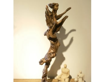 May include: A wooden sculpture of a figure with wings, standing on a square base. The sculpture is made from a natural piece of wood with a brown and gray color.