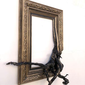 May include: An antique-style picture frame in gold tones, with a black inner border. A dark, root-like sculpture is integrated into the frame. The frame is mounted on a white wall.