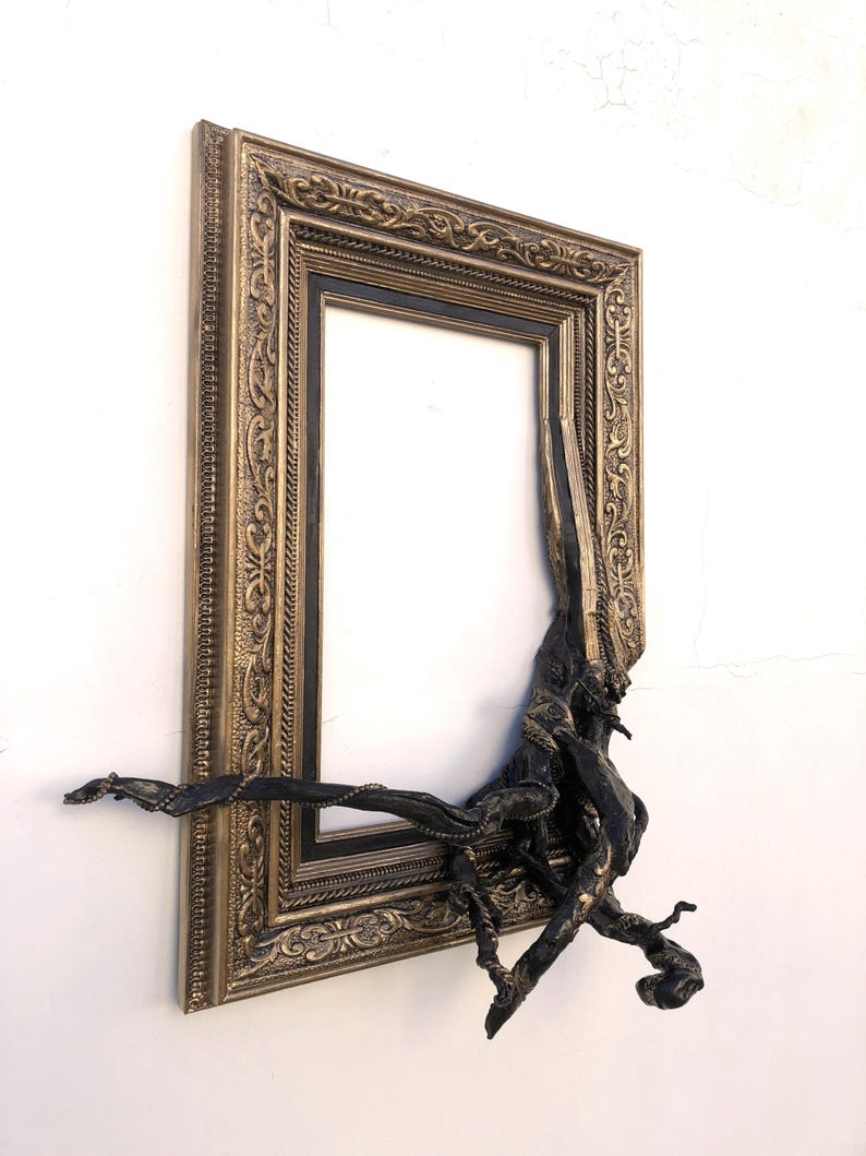 May include: An antique-style picture frame in gold tones, with a black inner border. A dark, root-like sculpture is integrated into the frame. The frame is mounted on a white wall.