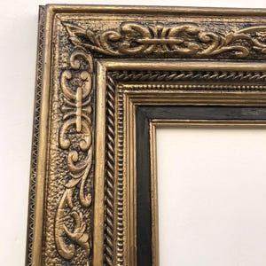 May include: An ornate gold picture frame with a black inner frame. The frame has detailed relief carvings and a textured finish. The design includes scrolling patterns and a rope-like border, creating a classic, elegant look. The frame is empty, ready for artwork or a photograph.