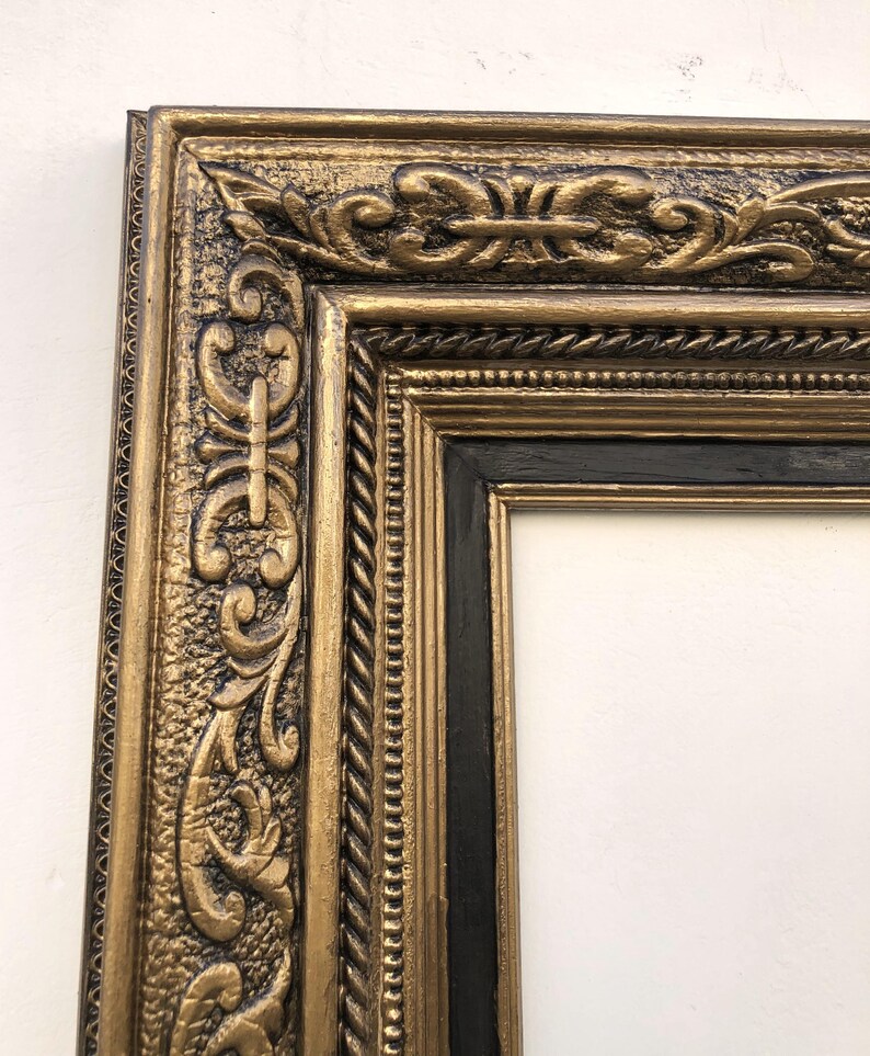 May include: An ornate gold picture frame with a black inner frame. The frame has detailed relief carvings and a textured finish. The design includes scrolling patterns and a rope-like border, creating a classic, elegant look. The frame is empty, ready for artwork or a photograph.