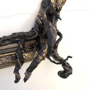 May include: An antique-style picture frame in dark bronze with gold detailing. The frame has elaborate carvings and a textured, organic appearance, with a hand or branch-like feature integrated into the frame's design.