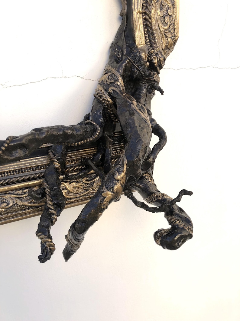 May include: An antique-style picture frame in dark bronze with gold detailing. The frame has elaborate carvings and a textured, organic appearance, with a hand or branch-like feature integrated into the frame's design.