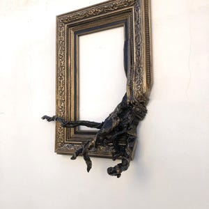 May include: An antique-style gold picture frame with intricate detailing, featuring a dark, textured sculptural addition. The frame is mounted on a white wall. The dark element appears to be emerging from the frame, adding a unique artistic touch.