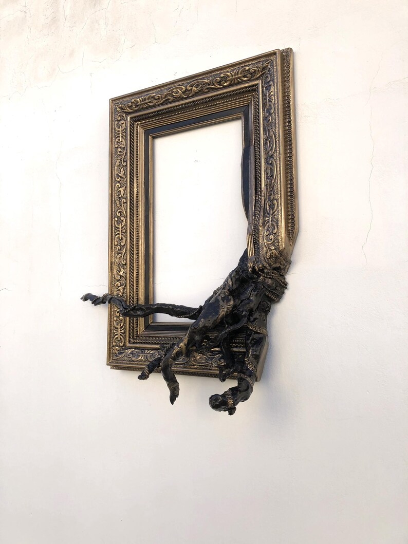 May include: An antique-style gold picture frame with intricate detailing, featuring a dark, textured sculptural addition. The frame is mounted on a white wall. The dark element appears to be emerging from the frame, adding a unique artistic touch.
