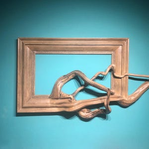 Może przedstawiać: Drewniana rama z naturalnym wykończeniem, z unikalną, organiczną rzeźbą przypominającą dryfujące drewno, która wydaje się wychodzić z ramy. Rzeźba jest jasnobrązowa i ma naturalną, szorstką fakturę.