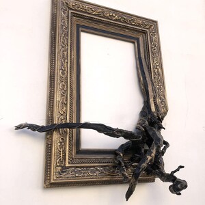May include: An ornate, rectangular picture frame with a black interior and a bronze-coloured, detailed border. A dark, organic sculpture appears to be growing out of the frame, adding a unique, artistic element to the design.