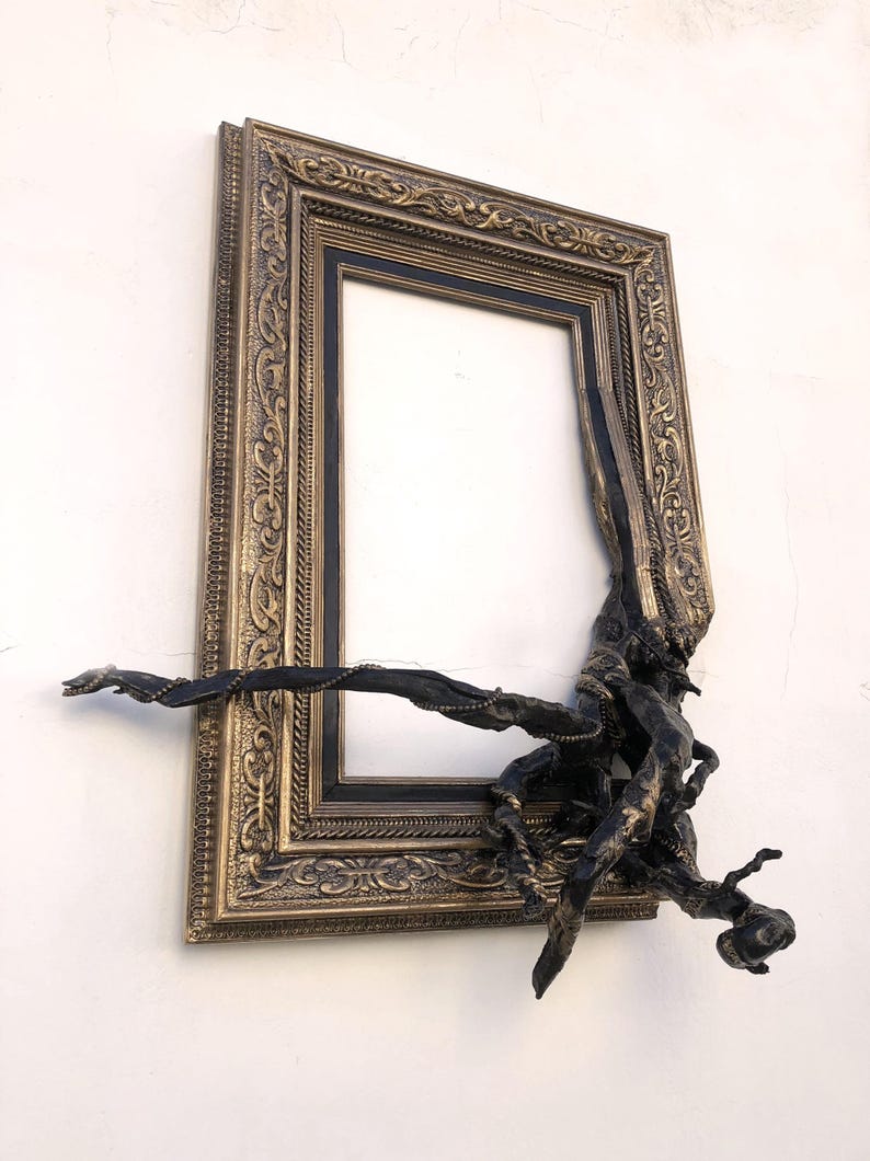May include: An ornate, rectangular picture frame with a black interior and a bronze-coloured, detailed border. A dark, organic sculpture appears to be growing out of the frame, adding a unique, artistic element to the design.