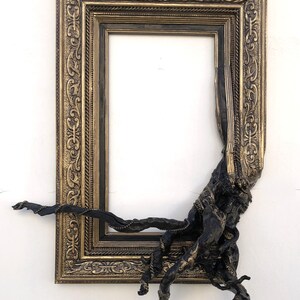 May include: An ornate gold and black picture frame with detailed patterns. A dark, textured sculptural element protrudes from the frame's corner, creating a unique artistic effect. The frame is empty, ready for a custom piece.