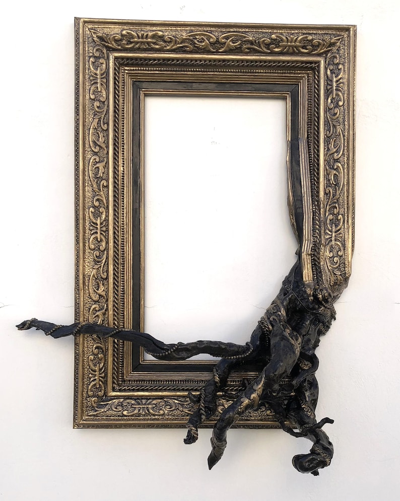May include: An ornate gold and black picture frame with detailed patterns. A dark, textured sculptural element protrudes from the frame's corner, creating a unique artistic effect. The frame is empty, ready for a custom piece.