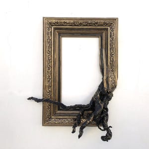 May include: An antique gold picture frame with a dark, textured, organic feature extending from the lower right. The frame has detailed carvings and a vintage aesthetic, set against a white backdrop.