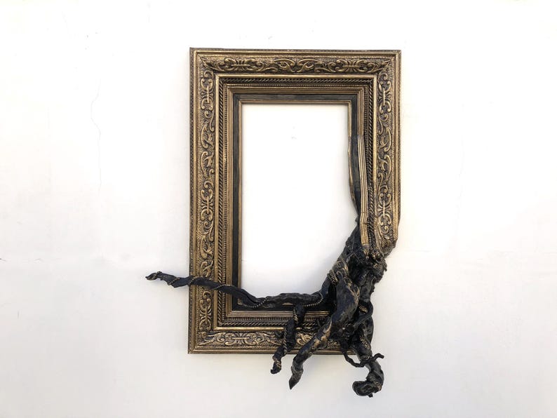 May include: An antique gold picture frame with a dark, textured, organic feature extending from the lower right. The frame has detailed carvings and a vintage aesthetic, set against a white backdrop.
