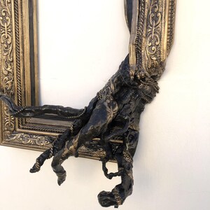 May include: An ornate gold and black picture frame with a sculpted, dark-coloured arm emerging from the frame. The frame has detailed carvings and a distressed finish, creating a unique, artistic piece.