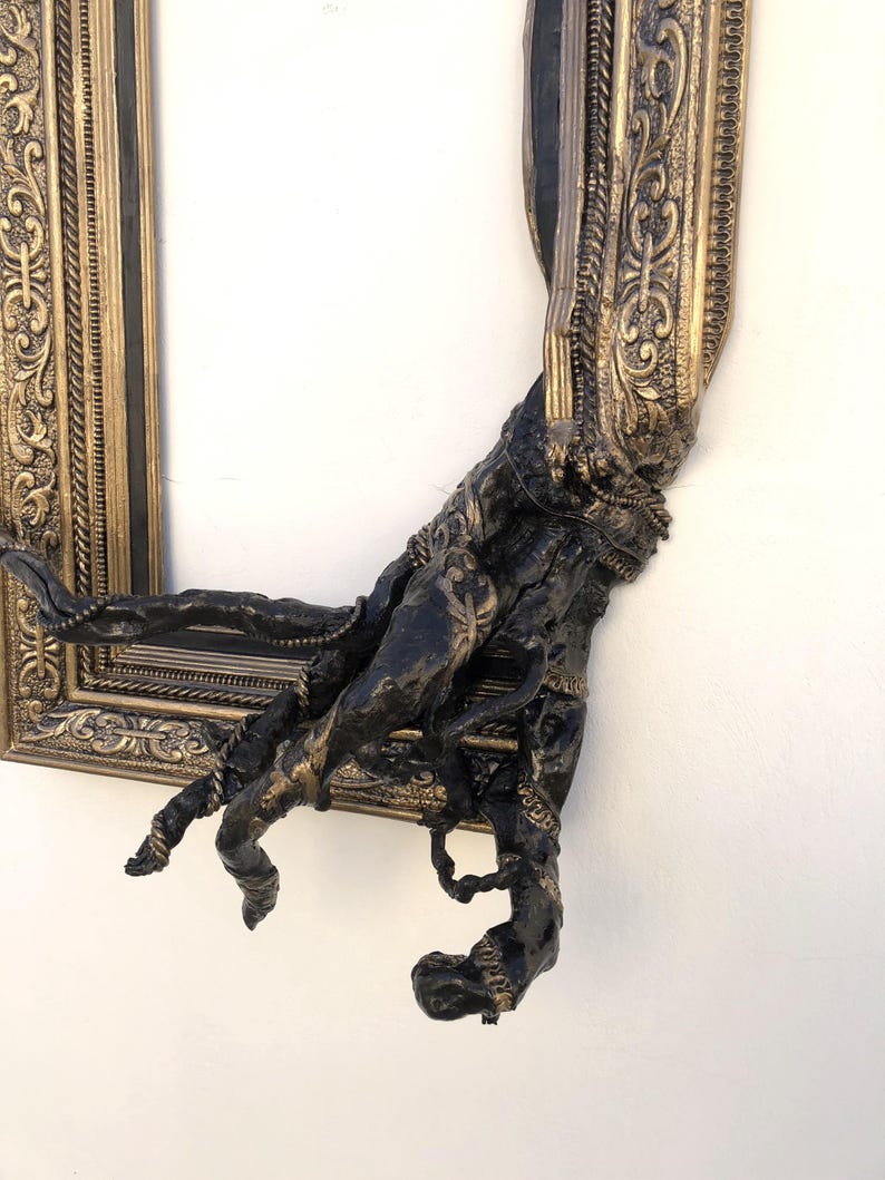 May include: An ornate gold and black picture frame with a sculpted, dark-coloured arm emerging from the frame. The frame has detailed carvings and a distressed finish, creating a unique, artistic piece.