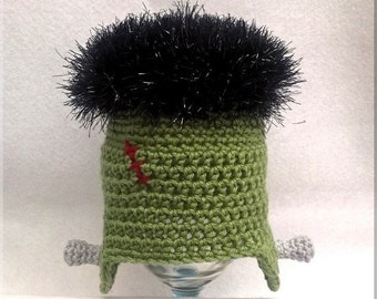 May include: Green crocheted hat with black fuzzy hair and gray crocheted arms. The hat has a red stitch on the side.  Ruffle Stitch Kids is written on the bottom of the image.