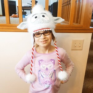 K&ouml;nnte beinhalten: Ein Kind tr&auml;gt eine wei&szlig;e und blaue H&auml;kelm&uuml;tze mit roten und wei&szlig;en Streifen und einem flauschigen wei&szlig;en Bommel. Die M&uuml;tze hat ein Monstergesicht aus Karikatur mit gro&szlig;en Augen und einem grinsenden Gebiss. Das Kind tr&auml;gt ein rosa Langarmshirt mit einem Eulenmotiv.