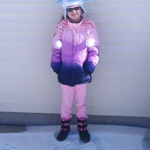 K&ouml;nnte beinhalten: Eine junge Person tr&auml;gt eine rosafarbene und violette Ombre-Winterjacke mit einer wei&szlig;en, flauschigen Schafkappe und schwarzen Stiefeln. Die Person steht im Schnee.