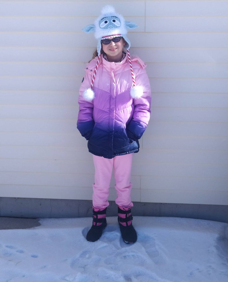 K&ouml;nnte beinhalten: Eine junge Person tr&auml;gt eine rosafarbene und violette Ombre-Winterjacke mit einer wei&szlig;en, flauschigen Schafkappe und schwarzen Stiefeln. Die Person steht im Schnee.