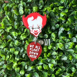 May include: Red and white glitter badge holder featuring a cartoon image of Pennywise the clown from the movie "IT" with the text "We all float down here" on a red heart-shaped charm.