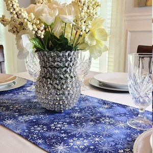 Peut inclure: Un chemin de table à motif flocon de neige blanc et bleu avec un vase en cristal transparent rempli de fleurs blanches et de baies blanches. Le vase est posé sur le chemin de table.