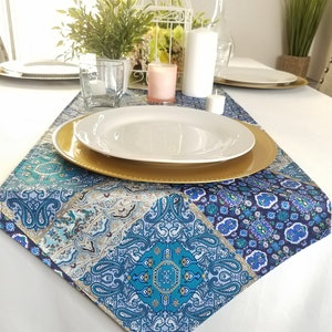May include: A blue and gold patterned table runner with a white plate and gold charger on top. The runner is on a white tablecloth. There are candles and a birdcage in the background.