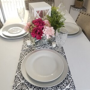 May include: A white table setting with a black and white damask table runner. The centerpiece is a white lantern with pink and white flowers and greenery. There are two white plates, two wine glasses, and two clear glasses on the table.