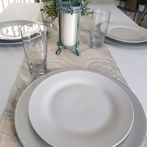 Può includere: Un piatto bianco per la cena con un piatto da portata argentato su un runner da tavolo in marmo. Ci sono due bicchieri trasparenti e un portacandele turchese con una candela bianca sul tavolo.