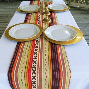 Peut inclure: Une nappe blanche avec un chemin de table rayé coloré. Quatre assiettes de service dorées avec des assiettes plates blanches sont disposées sur le chemin de table. Un bougeoir doré avec une bougie en verre transparent se trouve au centre du chemin de table.