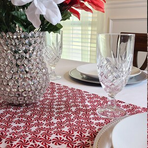 May include: A white tablecloth with a red and white peppermint swirl table runner. A clear glass vase filled with white and red poinsettia flowers sits on the table runner. Two clear glass wine glasses and a white dinner plate with a silver charger are on the table.