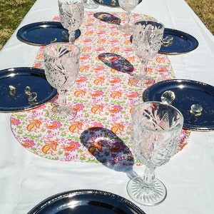 Pode incluir: Uma toalha de mesa branca com um corredor floral rosa e laranja. Seis copos de vinho de vidro transparente são dispostos no corredor, com pratos de prata colocados ao redor.