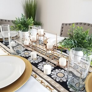 Peut inclure: Une table dressée avec une nappe blanche et un chemin de table à motifs noirs et dorés. La pièce maîtresse est un bougeoir en métal doré avec des bougies blanches. Il y a aussi des verres à eau en verre transparent et des assiettes de présentation dorées.