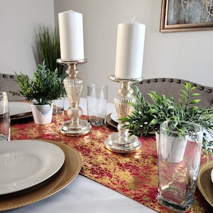 May include: A dining table set with two white pillar candles on decorative silver candlesticks. The table features gold chargers, white plates, and a red and gold patterned runner. Small potted plants and clear glasses add to the elegant arrangement.